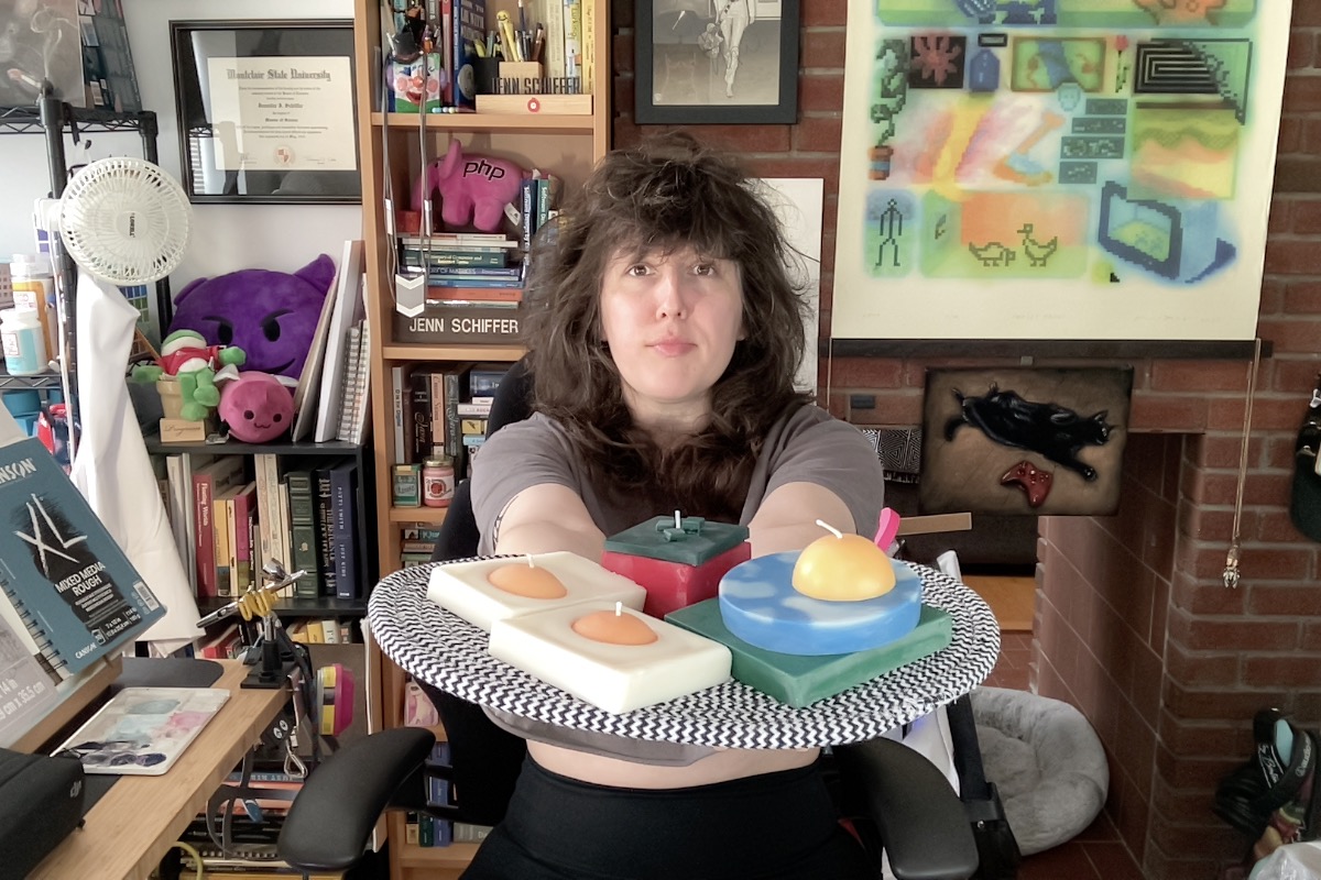 photo of me at my desk, today, and i'm holding up to the webcam a mat holding my new candles that i'm selling soon. they look like eggs, a tomato, and a outdoor scene