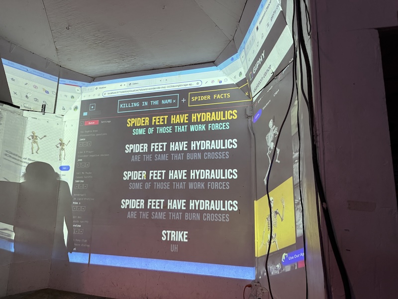 photo of the wonderville stage during robot karaoke, where someone is singing spider facts to the tune of killing in the name of. the lyrics repeating are "spider feet have hydraulics" in place of "some of those that work forces, are the same that burn crosses"