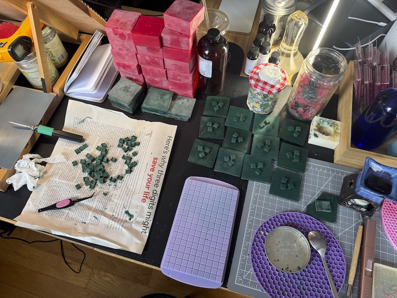 photo of my candle making space showing red blocks of wax, green tops with fake pixel leaves on them, the mold for the leaf pixels, a solder iron i use to melt stuff, and other tools.