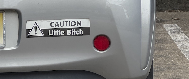 photo of the rear bumper of a car that has a sticker saying "CAUTION Little Bitch"