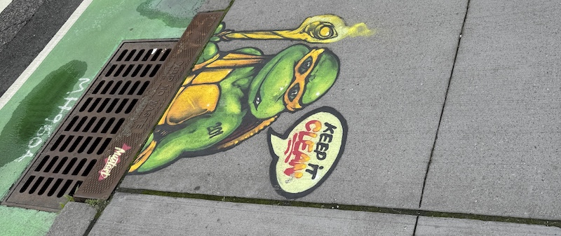 photo of the curb of a street near a gutter and someone painted michealangelo from teenage mutant ninja turtles above it saying "keep it clean"