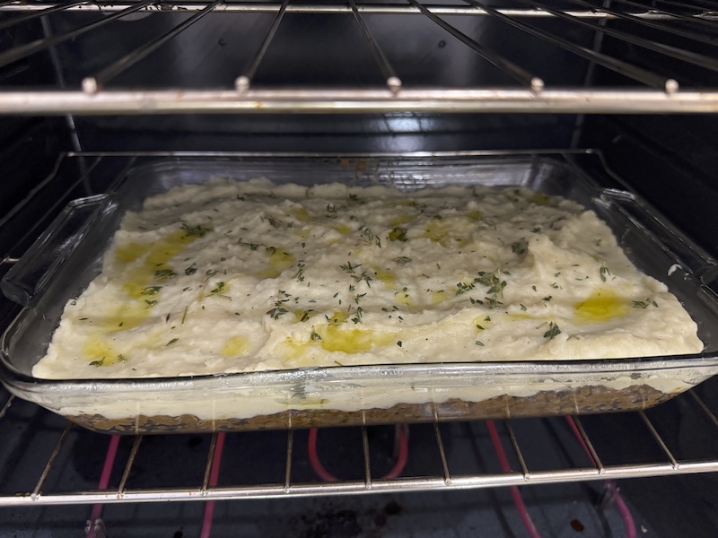 photo of my large glass baking dish of lentil shepherd's pie going into the oven, so the potatoes are mashed and cooked but there's no broiling yet. thyme and olive oil are spread on the top