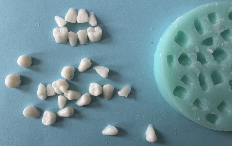 close up photo of a small silicone mold for making teeth shaped things, next to it are wax teeth i removed from the mold. there's 27 of them and they are small but i arranged a few to look like a tiny jaw