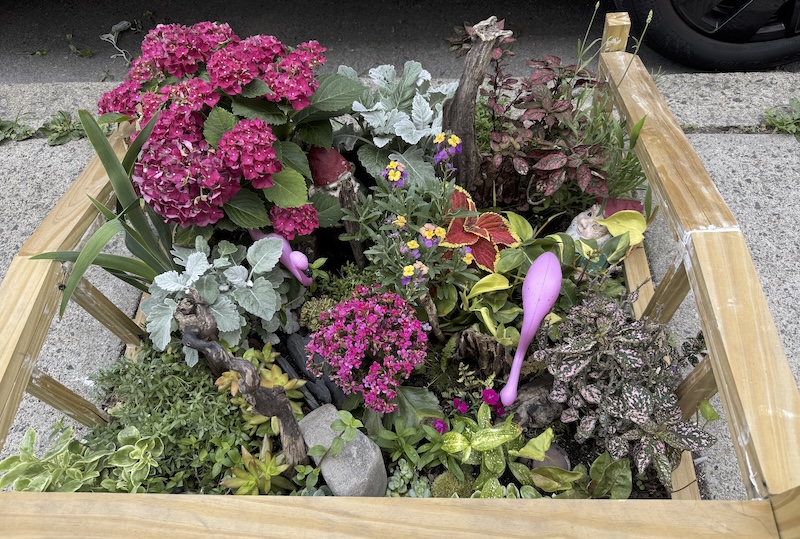 photo of a flower bed on the side of the road with a bunch of different plants and a gnome and a flamingo