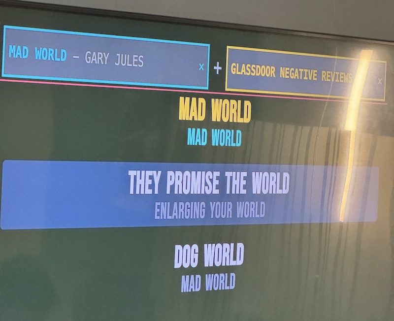 photo of a large classroom screen showing our robot karaoke interface. someone is singing "mad world" by gary jules but the lyrics come from glassdoor negative reviews. instead of the lyrics "mad world; enlarging you world; mad world" being sung the robot lyrics are "mad world; they promise the world; dog world"