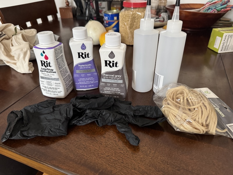 photo of my table with tie-dye supplies: black latext gloves, two empty squirt bottles, a bag of rubberbands, a bottle of rit colorstay dry fixative, bottle of hyacinth colored rit dye, and a bottle of charcoal gray colored rit dye