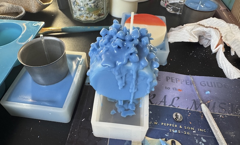 photo of a very messy candlemaking area where i've got one mold filled with blue wax and a votive mold inside it, another mold with a cylinder candle above it and i poured blue wax and crumbles onto it.