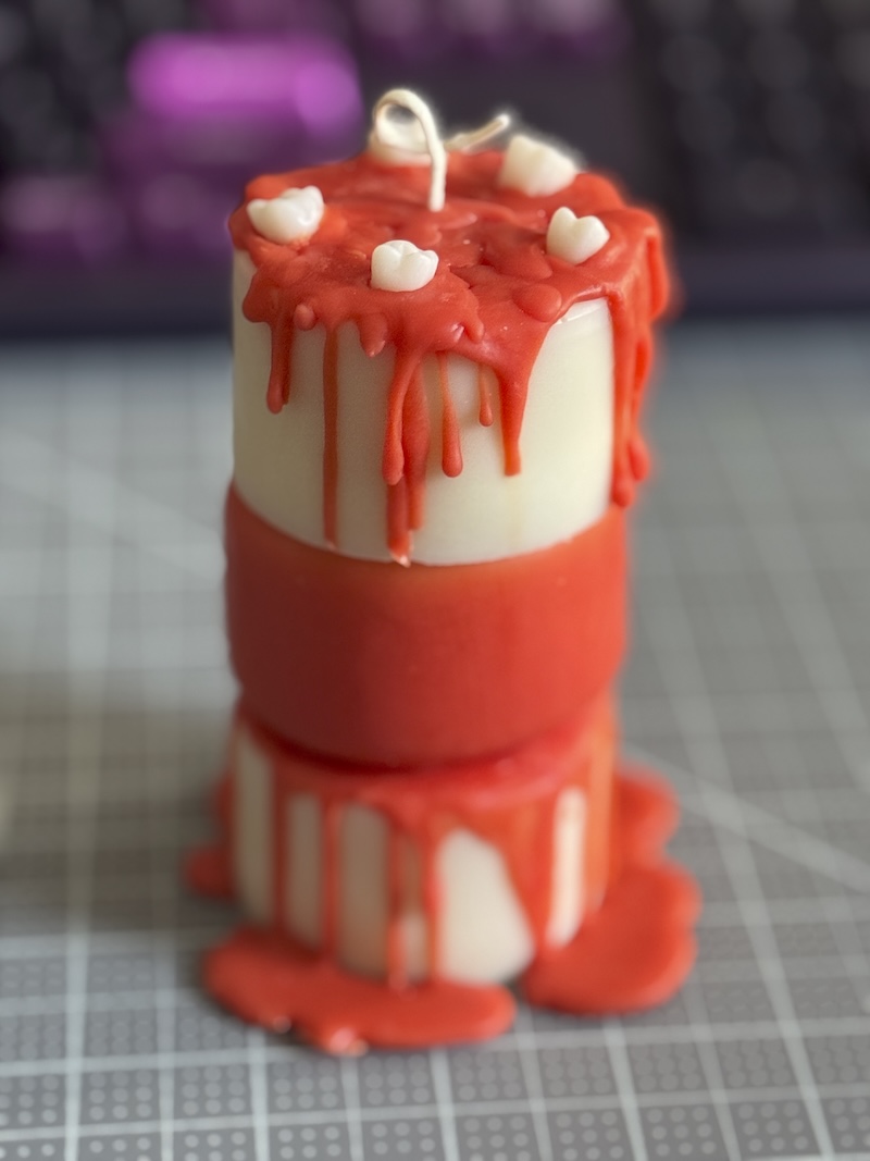 photo of a three tiered cylindrical candle I made. the bottom tier is white with a reddish orange wax dripped around it. The middle tier was dipped fully in that reddish orange wax and the top-tier has the reddish wax dripping and then there’s five wax white molars (teeth) decorating it. It’s like a fucked up bloody cake, which is just kind of how I was feeling in the dentist chair at the time. 