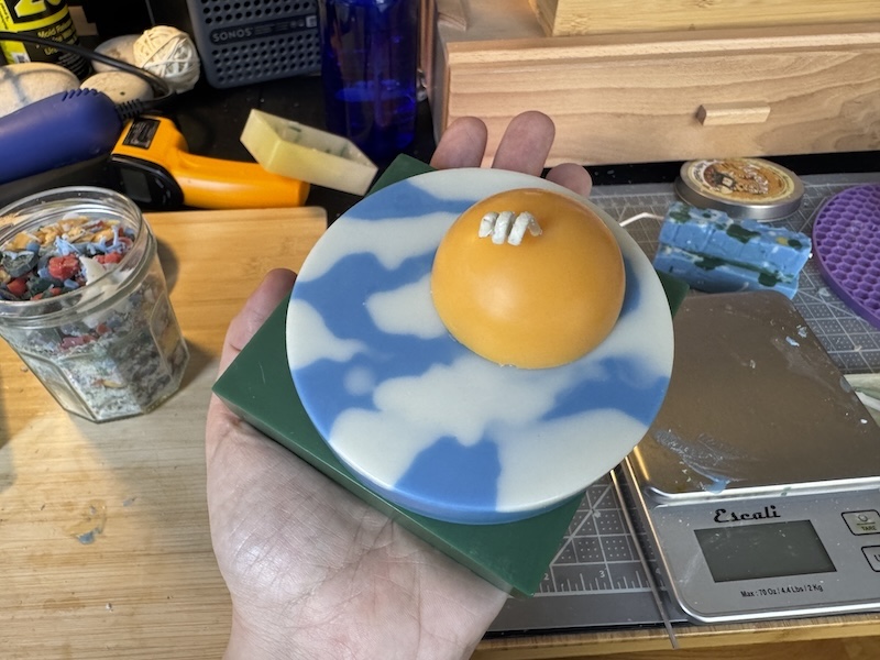 photo i took of my hand holding up one of my "touch grass" candles that has a green square bottom, a sky piece from the above photo, and a small yellow half-sphere as the sun on top. my candle-making space is messy lol