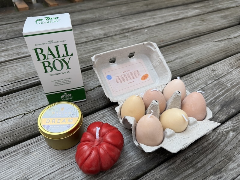 photo of a picnic table with 4 kinds of candles laid out: a box of the "ball boy" vacation candle, a "dream" scented gold tin from harlem candle company, a red heirloom tomato candle and a 6-pack carton of egg candles