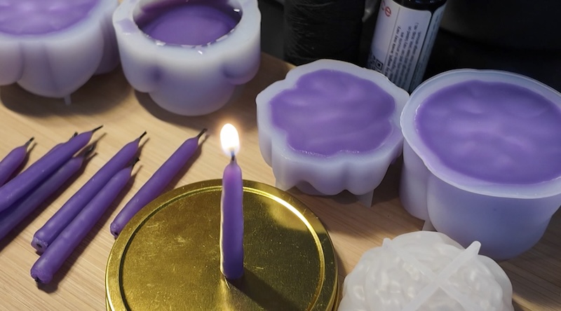 photo of my candle-making desk where I have a few hand dipped birthday candles that are “lilac” (purple) colored on the table, and there are some flower molds filled with the same purple wax and one of the candles I have stuck on a tin lid and it’s lit