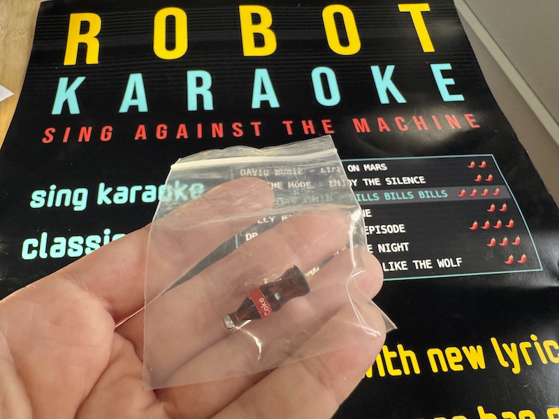 photo of a little baggie with a tiny coke bottle inside that i'm holding in front of a robot karaoke poster