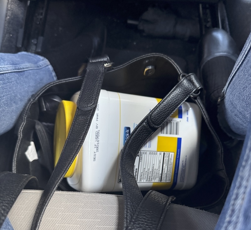 photo of my purse at my feet in a car and in the purse is a large plastic jug of liquid thickening powder