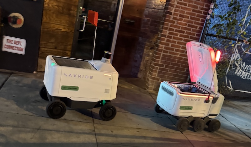 two white little delivery robots, one is open. they're both next to each other in front of one of the worst bars i've been to