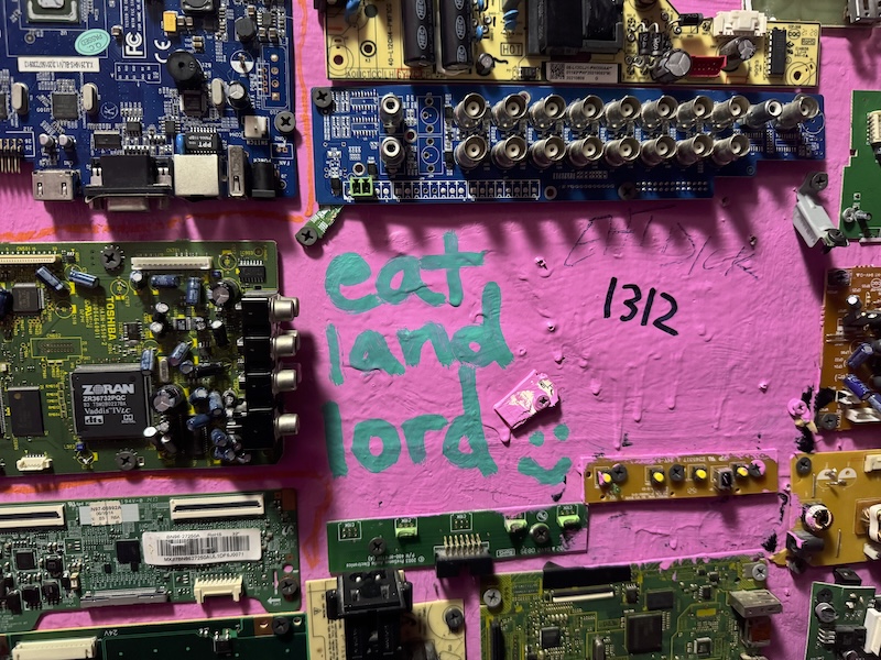 photo of a wall of a bathroom stall of wonderville. it's painted pink and with lots of computer board parts glued to it. someone wrote "eat land lord :)" and "1312" in marker