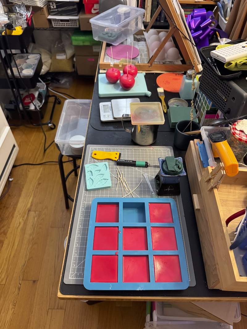 photo of my candle-making area, more than half of my L-shaped standing desk. i have dipped "apple candles" and some molds curing. it's organized and clean, but chaotic.