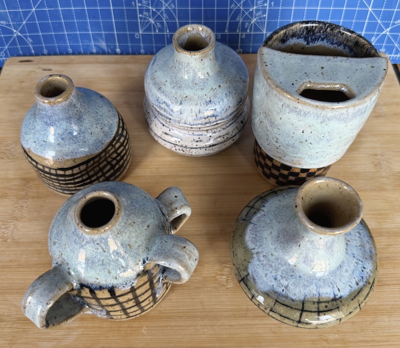 a photo of 4 bottles and a travel cup i made on the wheel, all with varying shapes but they have some kind of black grid, line or checker print and the top halfs are glazed witha periwinkle lavender glaze and the bottoms are either clear with the black lines or have some faint white glaze