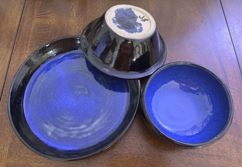 a photo of a blate and two bowls. the outside is black and the inside a cobalt blue. one of the bows are upside down to show how black the outside is and how dark the splotches of blue and black underglazes i used are