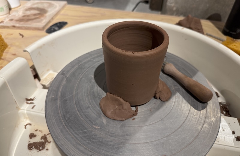 photo of a wheel in the clay studio and i'm trimming the top of a red clay cup that was thrown well because my classmate is really good and she made it