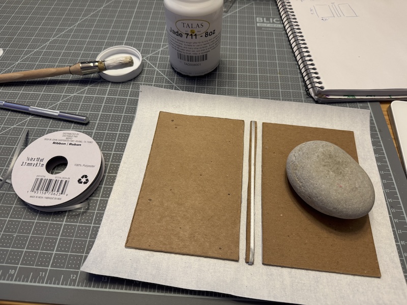 photo of my desk where i'm gluing book cover canvas onto some small notebook sized pieces of binding board. a rock is weighing down a glued piece