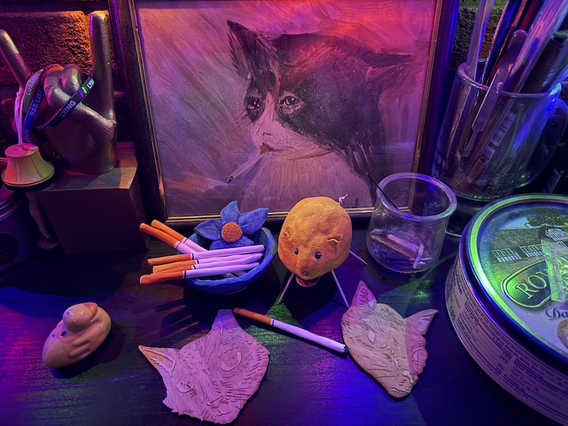 nighttime photo of a mantel at home showing my clay lemon pig next to some air dry clay and painted cigarettes. they are in front of a print of a painting of a cat smoking a cigarette, and behind some wooden coasters that are illustrations of pumagreg and jefrey (my late cat). you can also see a trophy and a cookie tin and incense