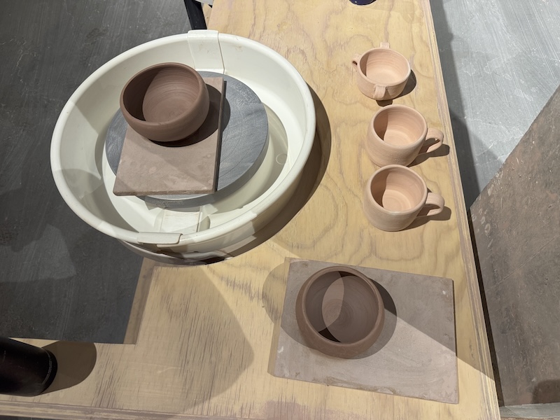 photo of my wheel setup at the ceramics studio, with three baked mugs and two bowls i'm in the process of trimming
