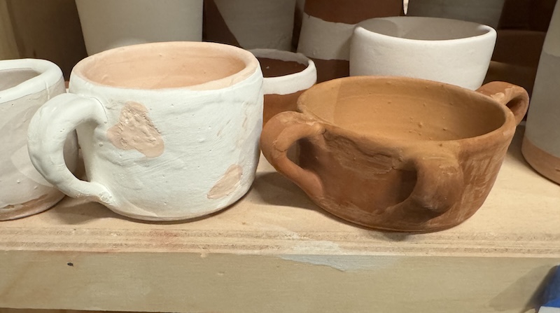 two sloppily glazed mugs on a shelf waiting to be fired