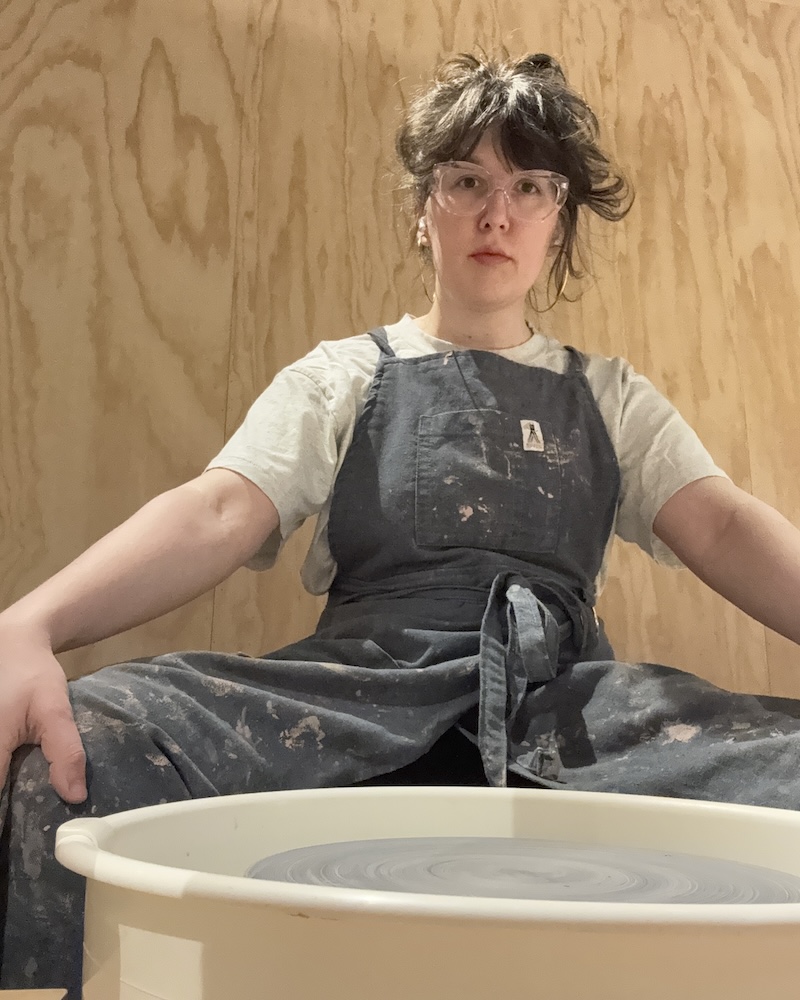 photo of me posing rigidly, seated in front of a clean pottery wheel with my hands on my knees, looking straight at the camera as i get ready to start a session of making absolute garbage out of clay