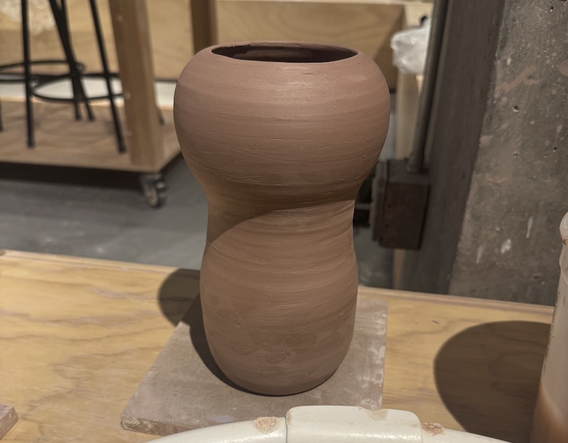 photo of a kinda tall vase that has a wider round top and more narrow long bottom, it looks like an upside down spaghetti squash now that I think about it. there’s a big hole on the top because it’s gonna be a vase. it’s just freshly trimmed so the clay is leather hard
