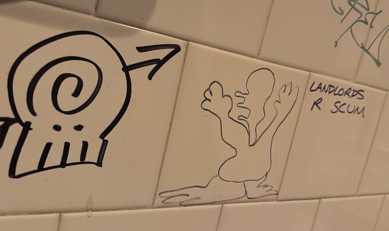photo of a few white tiles on the wall of the caveat bathroom. there's a skull illustration, an squiggly drawing of a guy, and next to that it says "LANDLORDS R SCUM"
