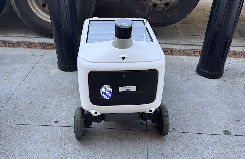 photo of a delivery robot on jersey city with a sticker on its face display that says "i steal jobs"