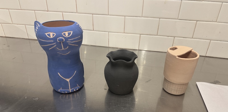 photo of three of my bisque pieces on a metal table in the studio. there's my blue cat vase, a black traditional round vase, and a travel mug that i drew a black grid on