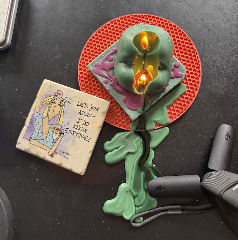 photo of a doll head candle i made melting with wicks in the head and out of the mouth and wax has flowed all onto my table. next to it i put a comic coaster that says "let's just assume i do know everything"