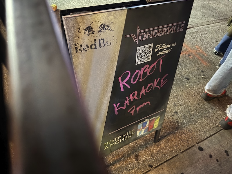 photo of a sandwich board outside and for wonderville that says "robot karaoke 7pm"
