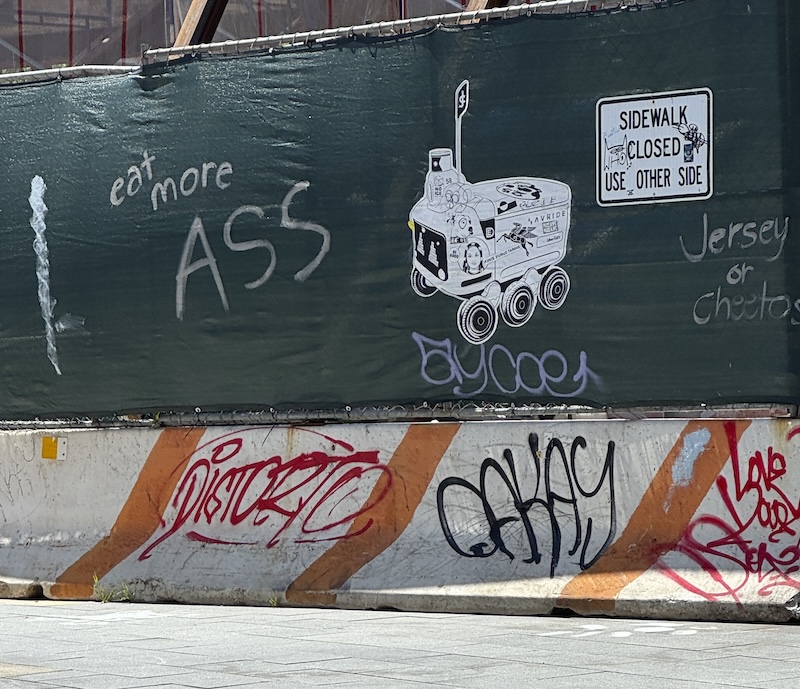 photo of a fence and road barrier around construction and someone freshly tagged it with "eat more ASS"