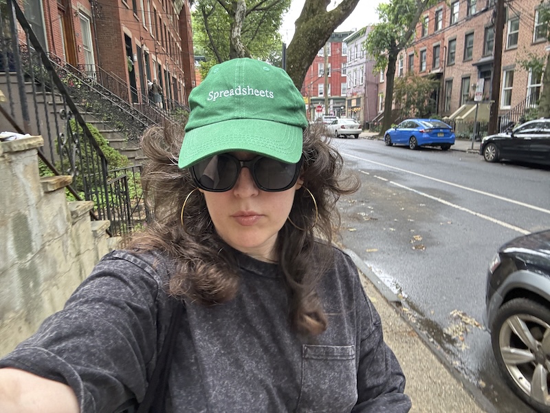 photo of me wearing a heavy oversized gray tee and a green hat that says "spreadsheets" outside in a humid and gray jersey city