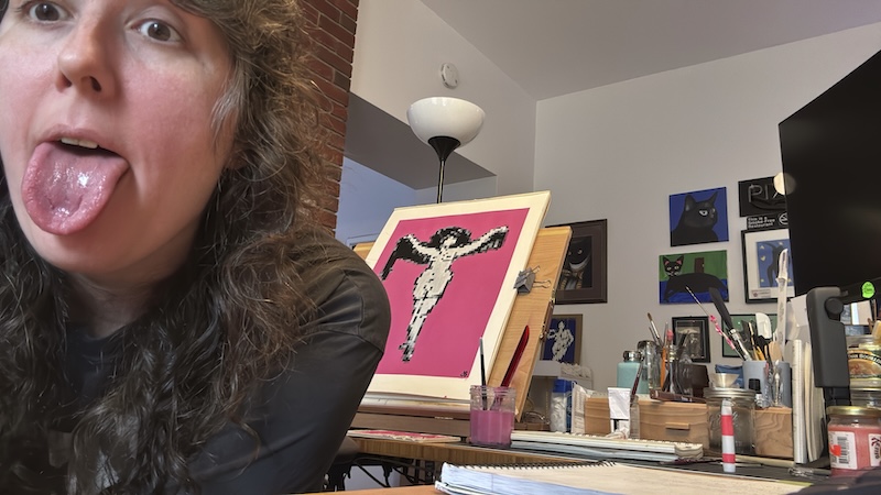 photo of me sticking my tongue out as i sit in front of and face away from a black and white with cosmos pink background pixel art gouache painting on my desk easel that i had just finished.