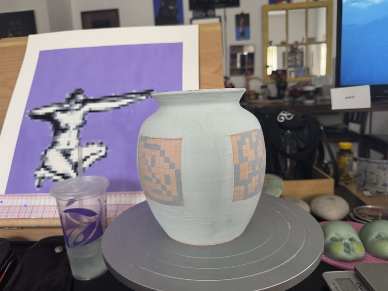 photo of a glazed vase with pixel art on it, sitting on a banding wheel on my desk near my recently finished pixel art figure painting