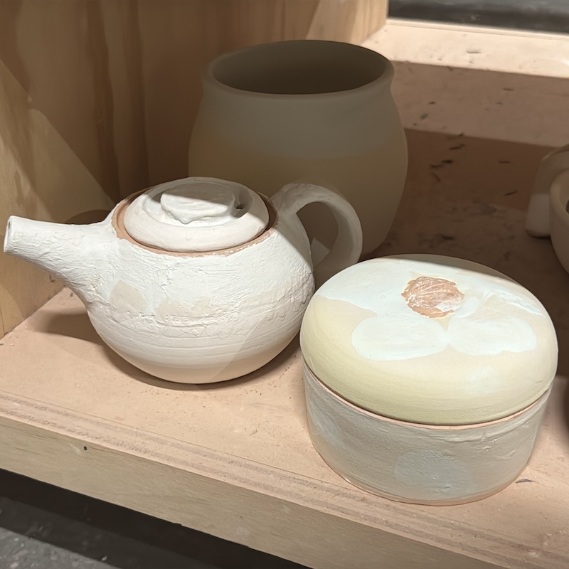A photo of three of my glazed pottery pieces on a shelf, ready to go into the kiln. In addition to my pot and enclosed vessel there’s a teapot that appears to have the same cream colored glaze around it with some white pasty spackle like glaze on the top half. you can see a whisper of horizontal lines underneath the cream glaze.
