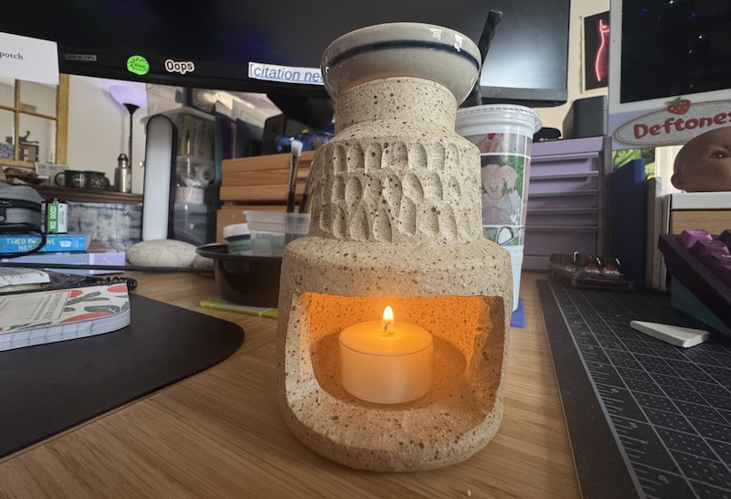 a photo of my unglazed wax melter with a small dish on top, and a tealight candle lit inside. it's on my desk at home.