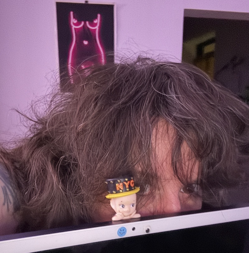 a photo of me hiding behind my computer monitor and on it peeking toward the camera with me is a sonny angel doll that has a top hat that says "NYC" on it