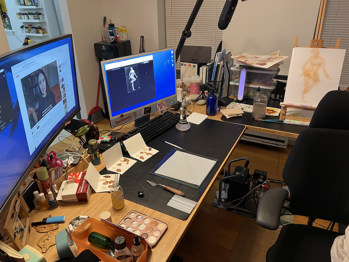 photo of my entire workspace, showing an L-shaped desk with a computer, large 2nd monitor, and an on-desk easel setup. the desk has a lot of stuff all on it, but it's organized