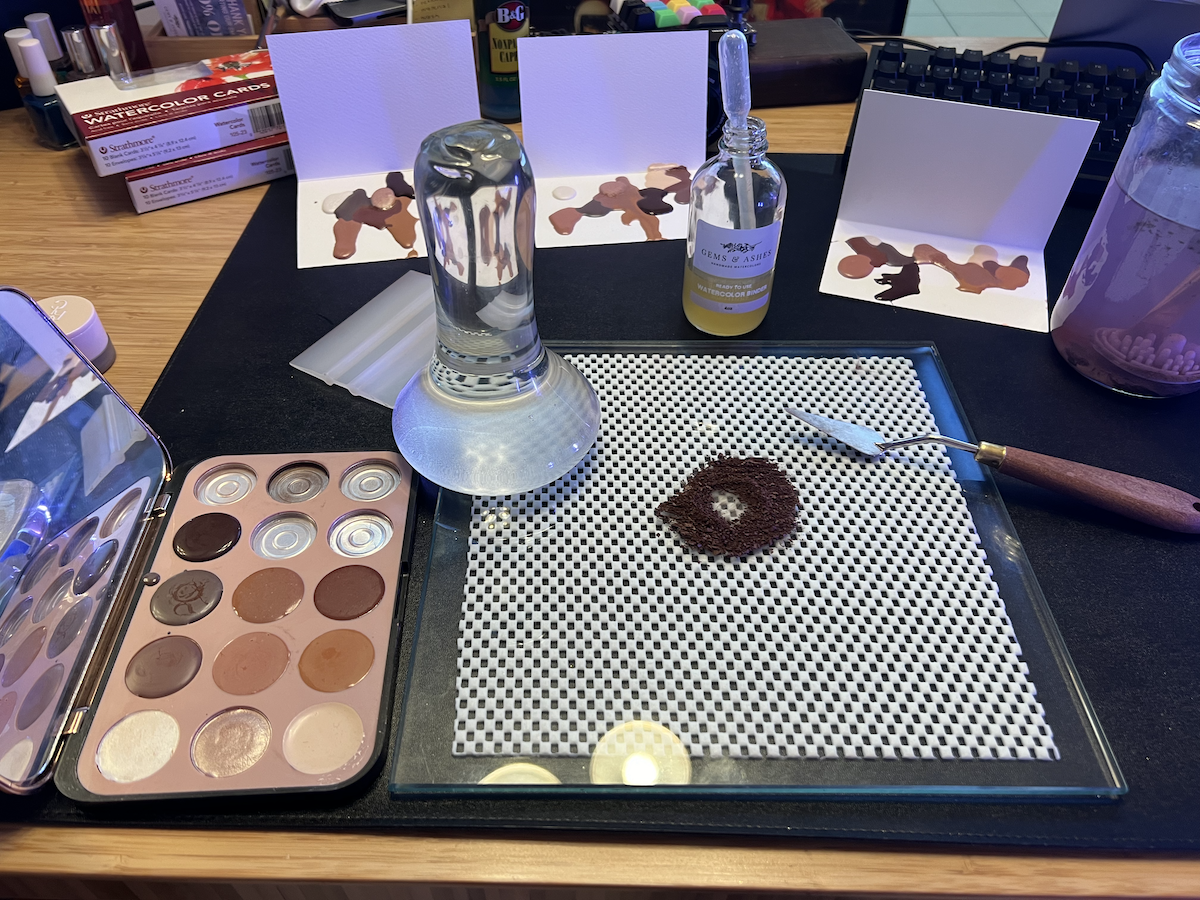photo of my watercolor-making workspace, including a glass slab with a small pile of brown pigment, a glass muller and the eyeshadow palette i'm working from