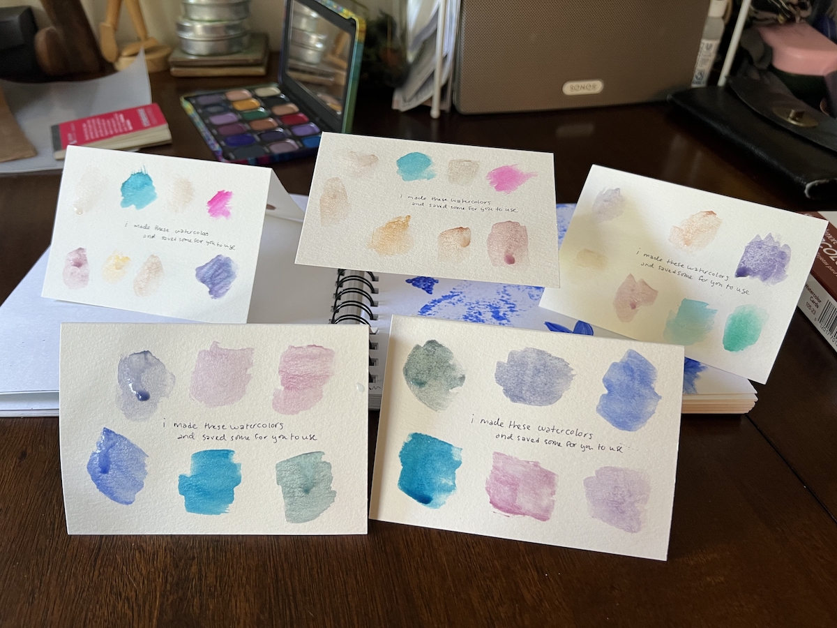 photo of my table with 5 watercolor paper cards spread out. each have swatches of the colors inside and they say 'i made these watercolors and saved some for you to use'"'