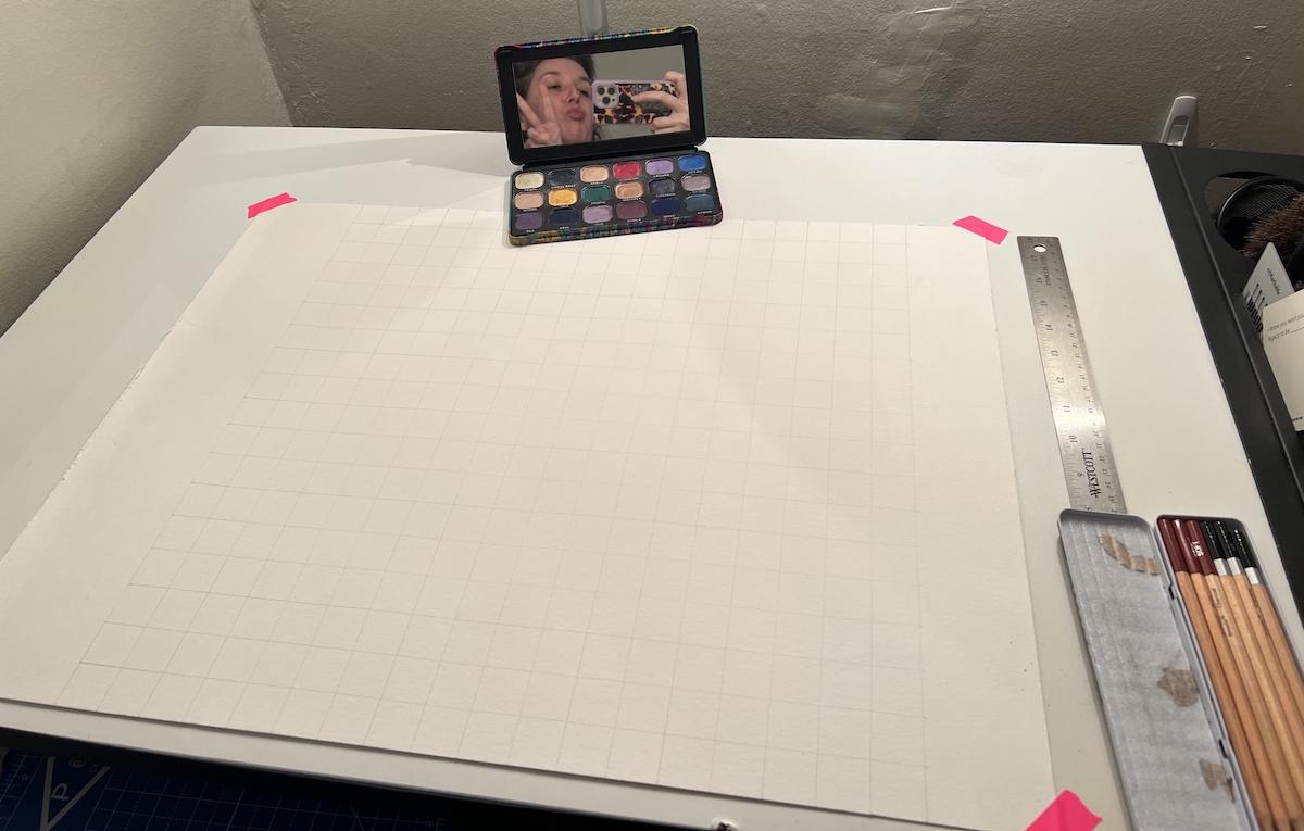 photo of my drafting table with a large watercolor paper that has a light grid of 18 by 18 squares on it in pencil. the palette is on the top and you can see me giving face in the mirror of the palette
