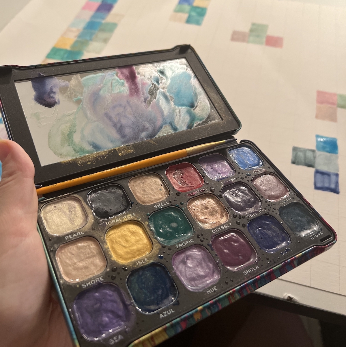 photo of my hand holding the used watercolor palette and a partially finished painting of a grid of colors from the palette in the background