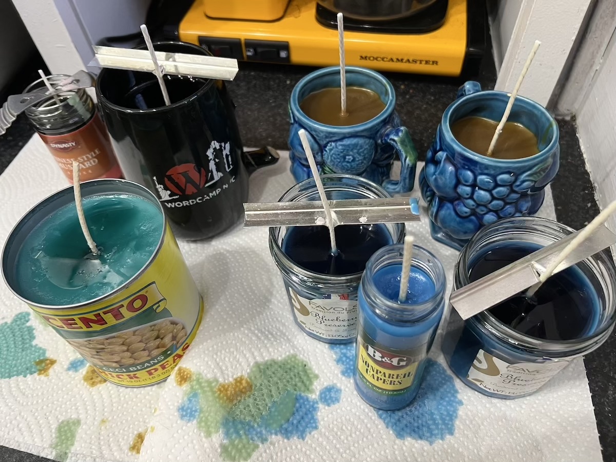 photo of in my kitchen of the candles i've poured and are curing, still mostly melted. there's a chinese mustard jar, 3 mugs, two jam jars, a chickpea can, and a capers jar
