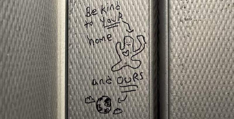 photo of a bathroom stall door where someone wrote "be kind to your home and ours" with a little guy and a planet earth, all drawn in sharpie
