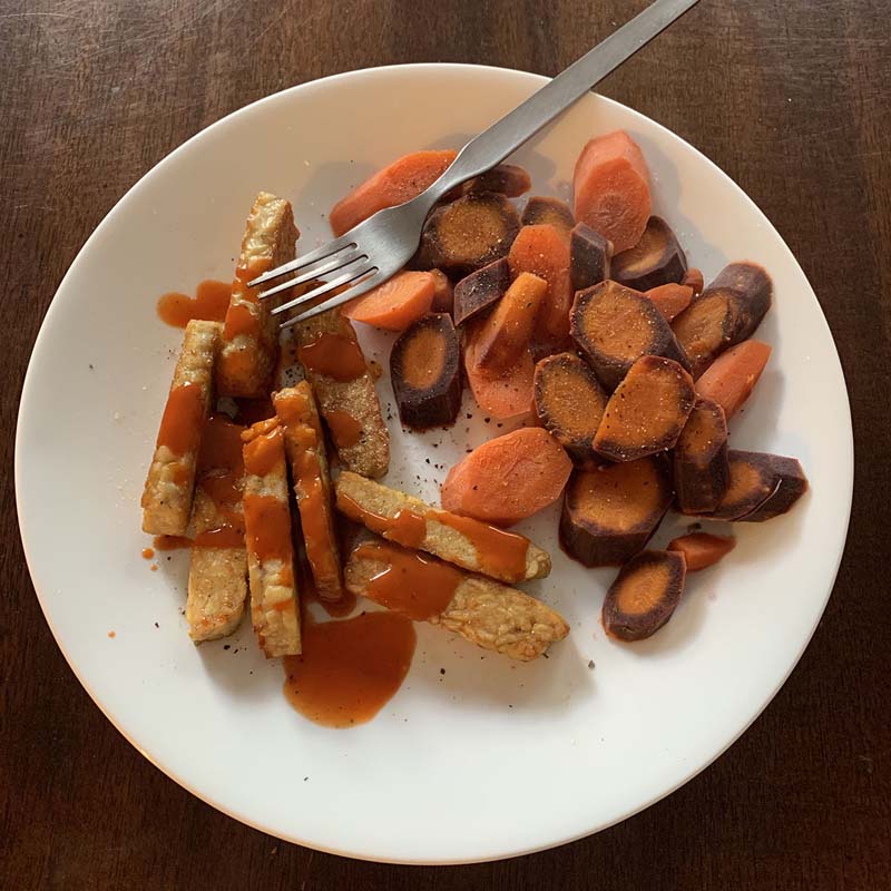 a plate with carrot coins and tempeh covered in red hot sauce