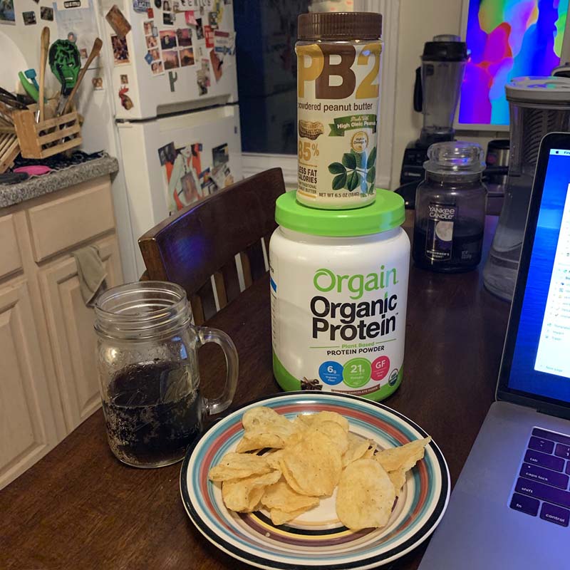 a glass of coke zero and a plate with chips in front of a cannister of protein powder and peanut butter powder